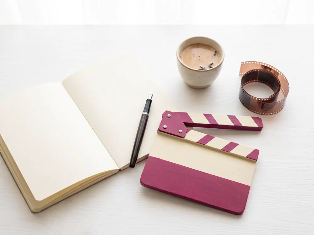 An open notebook with a fountain pen, a small clapperboard and a cup of chai on a white desk