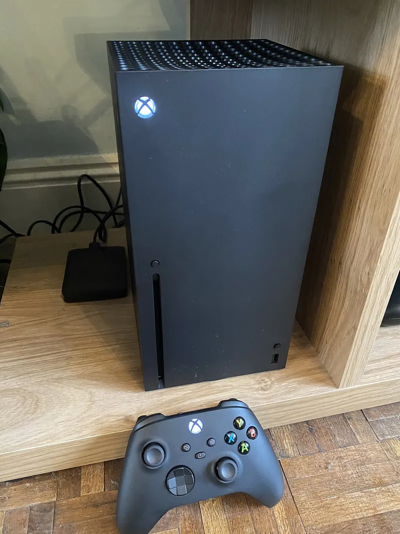 Black Xbox Series X console standing upright on a shelf with a controller in front