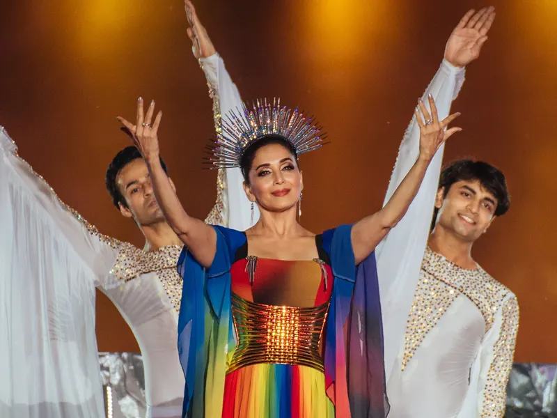 Madhuri Dixit smiling with both arms raised, wearing a jewelled halo headpiece and a rainbow gown, flanked by two dancers in white