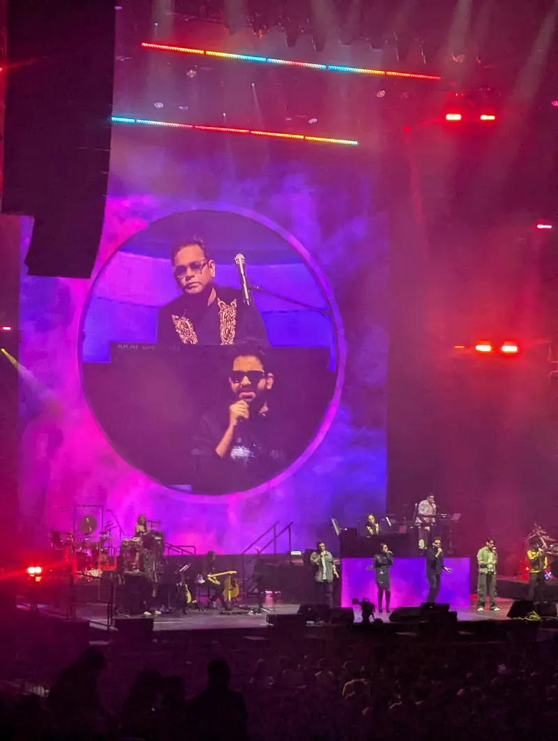 A concert stage with a large circular screen showing A.R. Rahman at a keyboard and A.R. Ameen singing