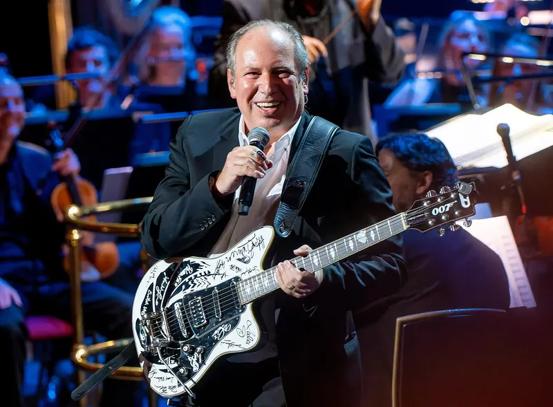 Hans Zimmer smiling with a microphone and an autographed electric guitar in front of an orchestra