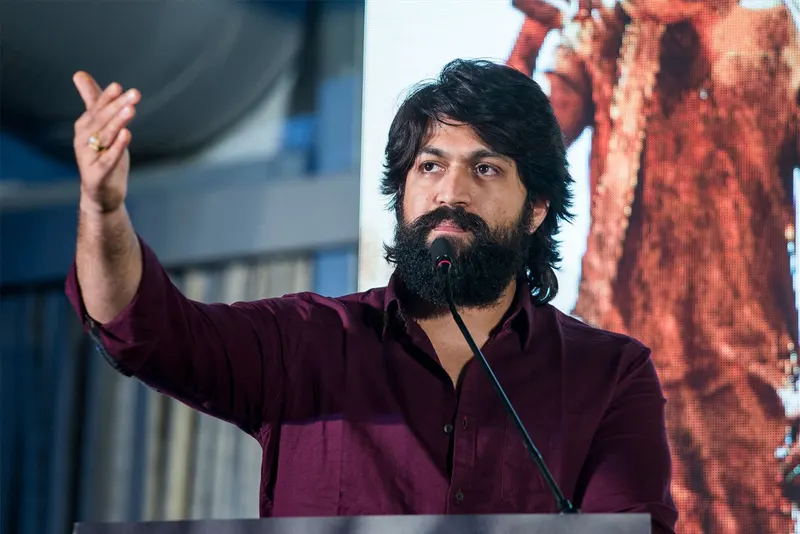 Yash in a maroon shirt gesturing as he speaks into a microphone