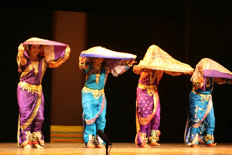 Four Lavani dancers in purple and blue nauvari saris holding their pallus over their heads on stage
