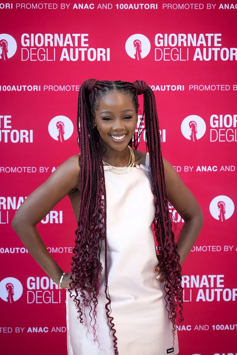 Thuso Mbedu in a white dress with long braids on a red backdrop