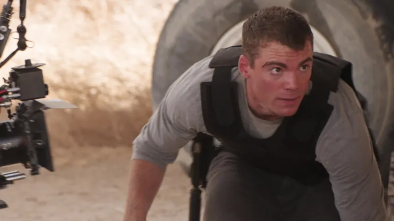 Gabriel Basso in a tactical vest crouching on a sandy film set beside a camera