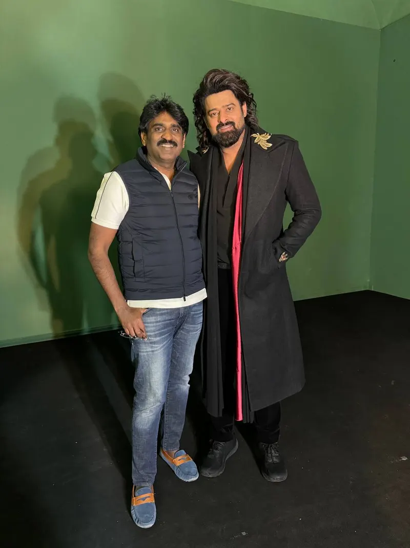 Prabhas in a long dark coat posing with a man in a studio