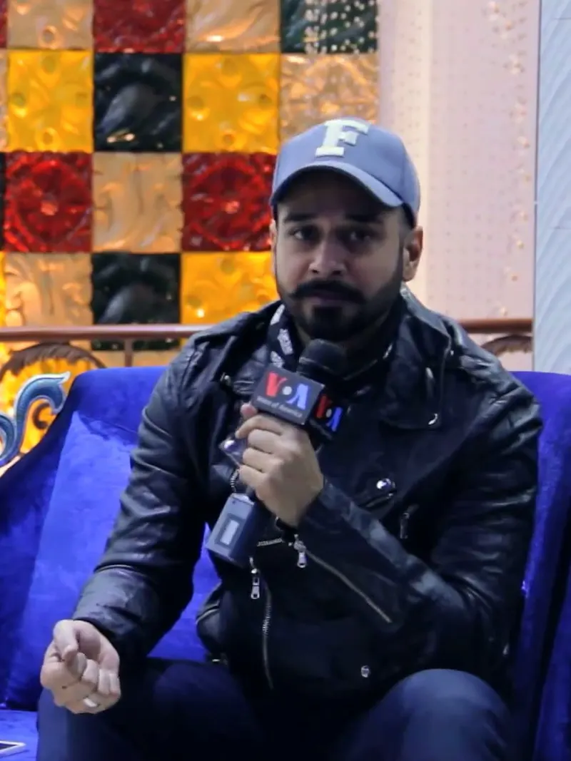 Faysal Quraishi in a cap and leather jacket holding a microphone