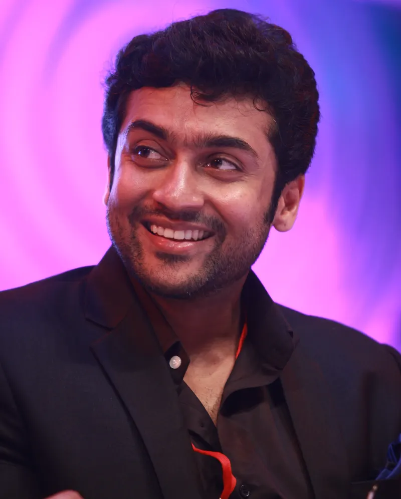 Suriya smiling on stage against a pink backdrop