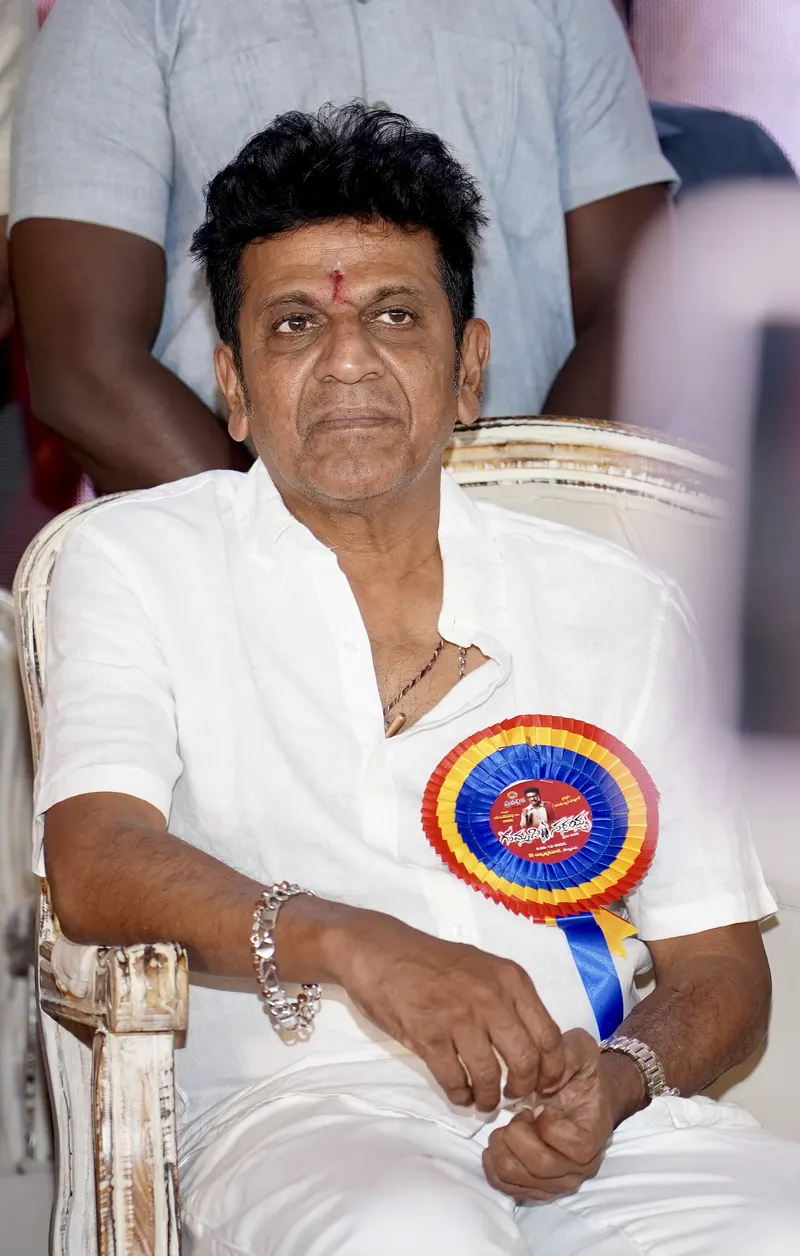 Shivarajkumar seated on stage wearing white with a rosette